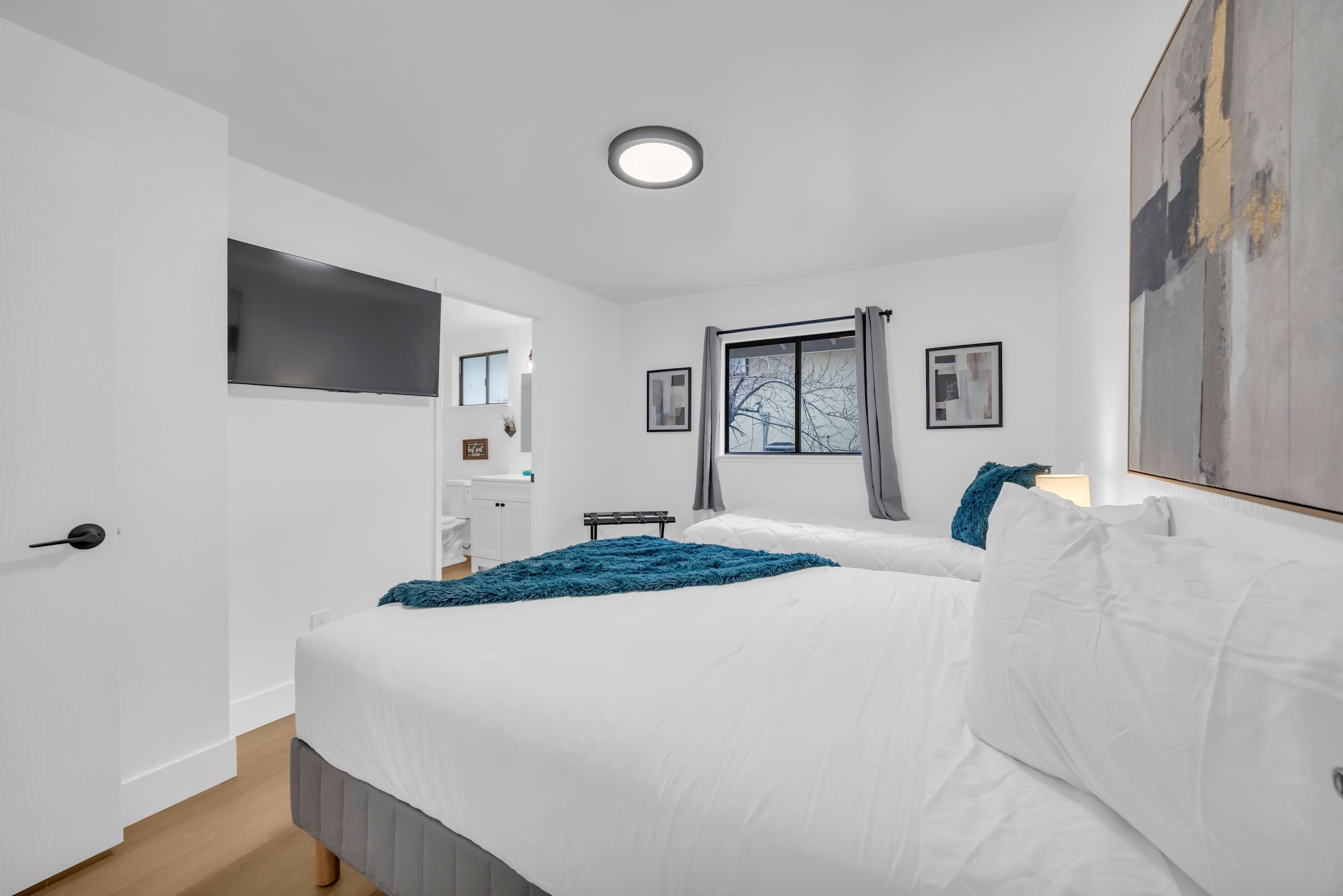 Een moderne slaapkamer met een groot bed, een flatscreen-televisie aan de muur, een raam met gordijnen en een schilderij.