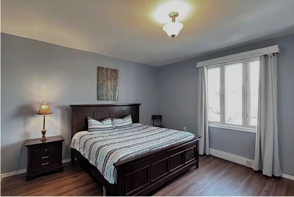 A bedroom with a large bed, a nightstand with a lamp, a ceiling light, a window with curtains, and a painting on the wall.