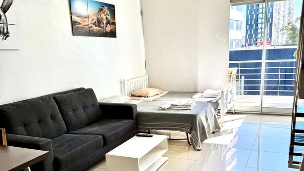 Ein modernes Wohnzimmer mit einem schwarzen Sofa, einem weißen Couchtisch und einem Bett mit Blick auf Gebäude durch ein großes Fenster.