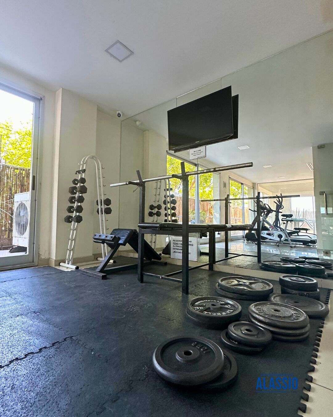 Un gimnasio con equipo de levantamiento de pesas, televisión y un espejo grande.