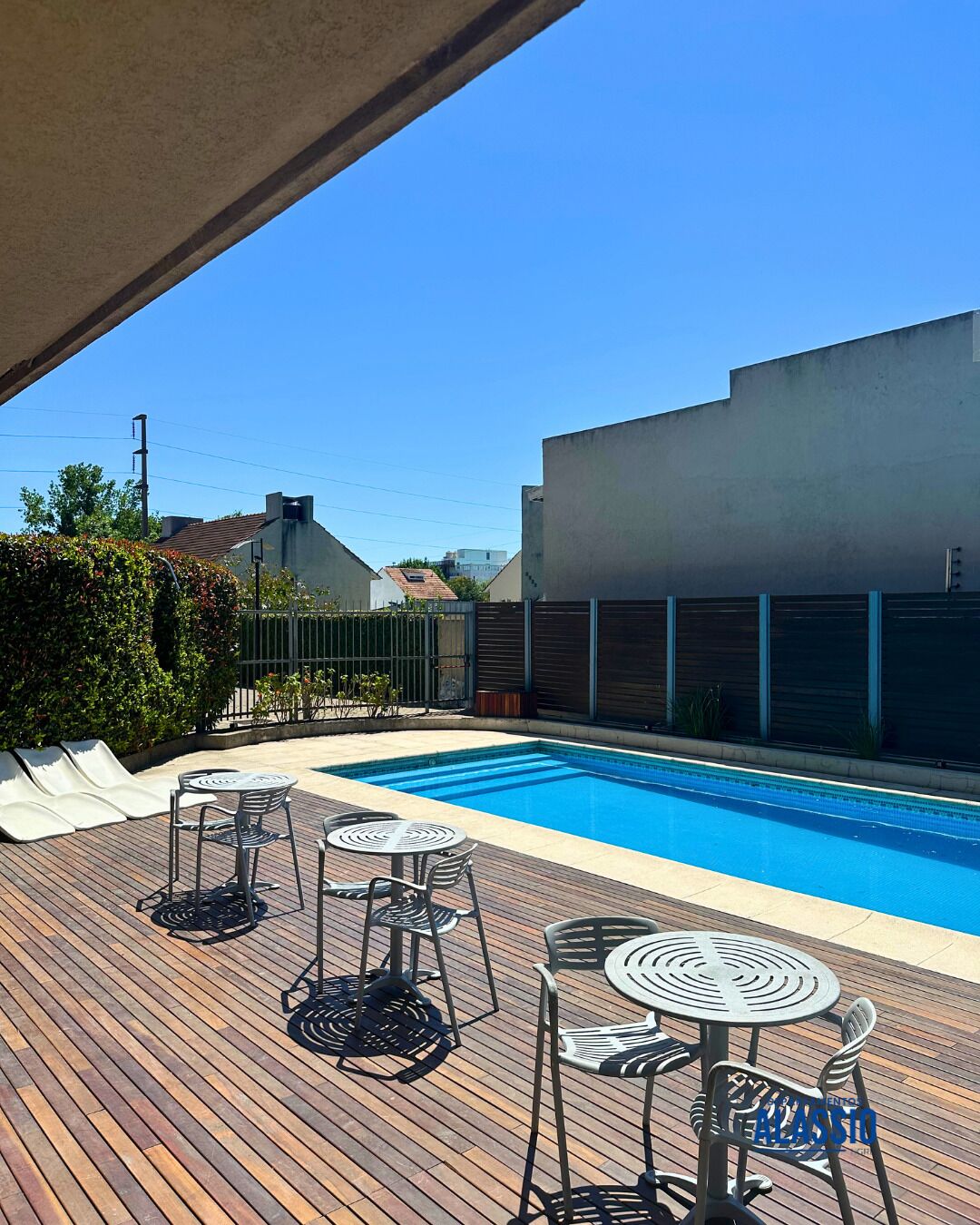 Una piscina con sillones y mesas en una terraza de madera, rodeada por una cerca y edificios.