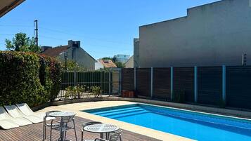 Una piscina con sillones y mesas en una terraza de madera, rodeada por una cerca y edificios.