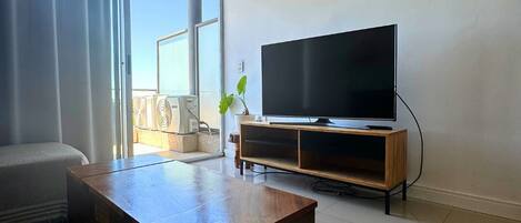 Ein Wohnzimmer mit einem Flachbildfernseher, einem Holztisch und einem großen Fenster mit Vorhängen.