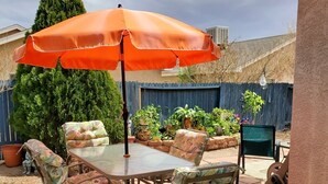Eine Terrasse im Freien mit Tisch und Stühlen, einem orangefarbenen Sonnenschirm und einem blauen Zaun im Hintergrund.
