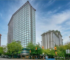 A tall, modern hotel with a glass facade, located at a city intersection with traffic lights and trees. - Bright 1 Bedroom Condo in the Heart of the City! (Vancouver)