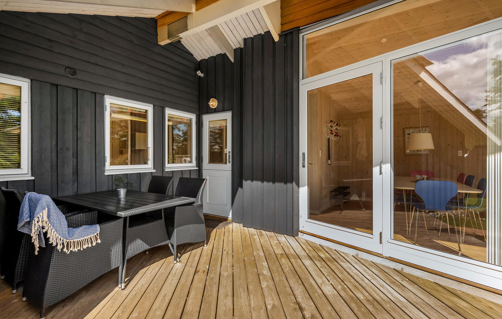 A wooden deck with a table and chairs, a sliding glass door leading to an indoor dining area, and a dark exterior wall.