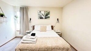 Ein Hotelzimmer mit Bett, Nachttischen, einer Nachttischlampe, einem Fenster mit Vorhängen und einem Wandbild.