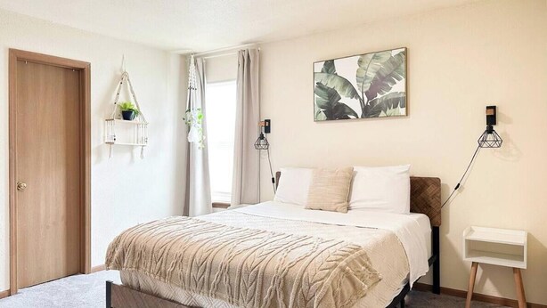 Ein Schlafzimmer mit einem Bett, einem Nachttisch, einer hängenden Pflanze, einer Wandlampe und einer Tür.