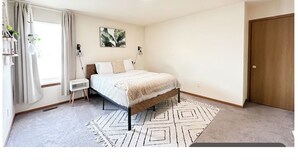 Ein Schlafzimmer mit Bett, Nachttisch, Vorhängen am Fenster, einer Wandlampe und einer Tür.