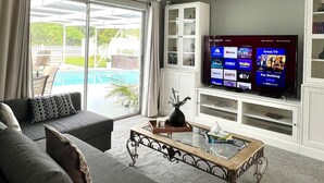 Ein Wohnzimmer mit einem großen Flachbildfernseher, einem Sofa, einem Couchtisch und Blick auf einen Swimmingpool durch große Schiebeglastüren.