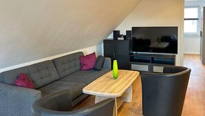 Un salon moderne avec un canapé d’angle gris, une table basse en bois, un fauteuil noir et un pouf noir. Un téléviseur à écran plat est posé sur un meuble noir, et une petite enceinte noire se trouve sur une commode noire.