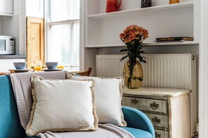 Ein gemütliches Wohnzimmer mit einem blauen Sofa, zwei weißen Kissen, einer Vase mit Blumen und einem Bücherregal mit Büchern und Dekoration.