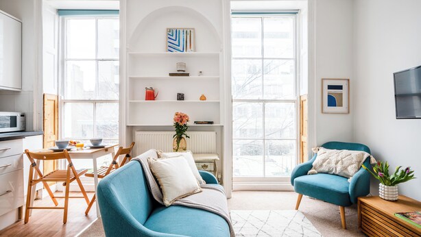 A modern living room with a blue sofa, a matching armchair, and a dining table with chairs. There is a bookshelf with books and decorative items, and a large window providing natural light. - The Russell Collection (London)