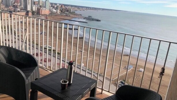 Een balkon met rieten meubels, een metalen hekwerk en uitzicht op het strand en de stad.