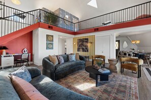 A spacious living area with a large sofa, a coffee table, and a dining table. There is a staircase with a red railing leading to an upper level.