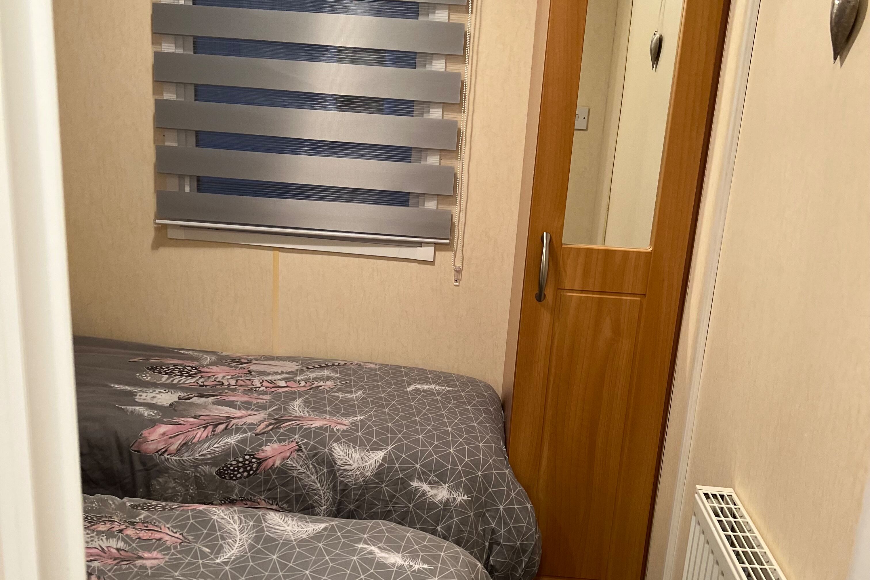 A narrow room with a bed featuring a geometric pattern and feather design, a window with horizontal blinds, and a wooden door.