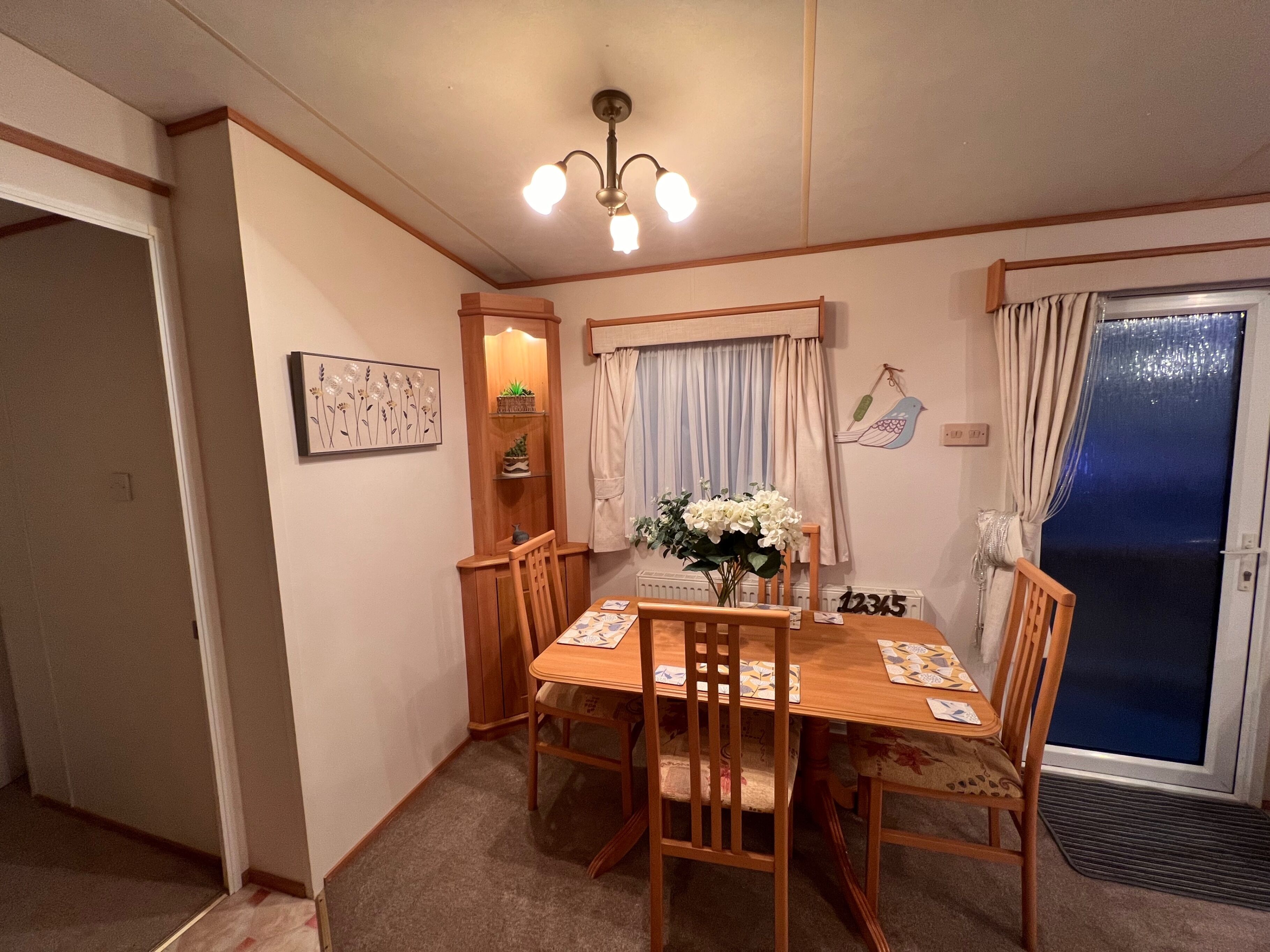 A dining area with a wooden table, four chairs, a chandelier, a window with curtains, a wall-mounted bird decoration, and a door with a blue screen.