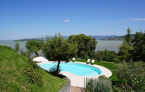 Ein Pool mit geschwungener Kante, umgeben von einer Rasenfläche und Bäumen, mit Blick auf einen See und sanfte Hügel in der Ferne.