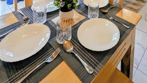 Un coin repas avec une table dressée pour quatre personnes, un canapé agrémenté de coussins à motifs, une cuisine en arrière-plan et une fenêtre avec des rideaux jaunes.