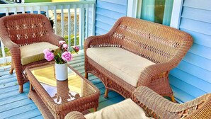 A wicker outdoor seating area with a glass-top table and chairs, a vase with flowers, and a view of a house with blue siding and a window.