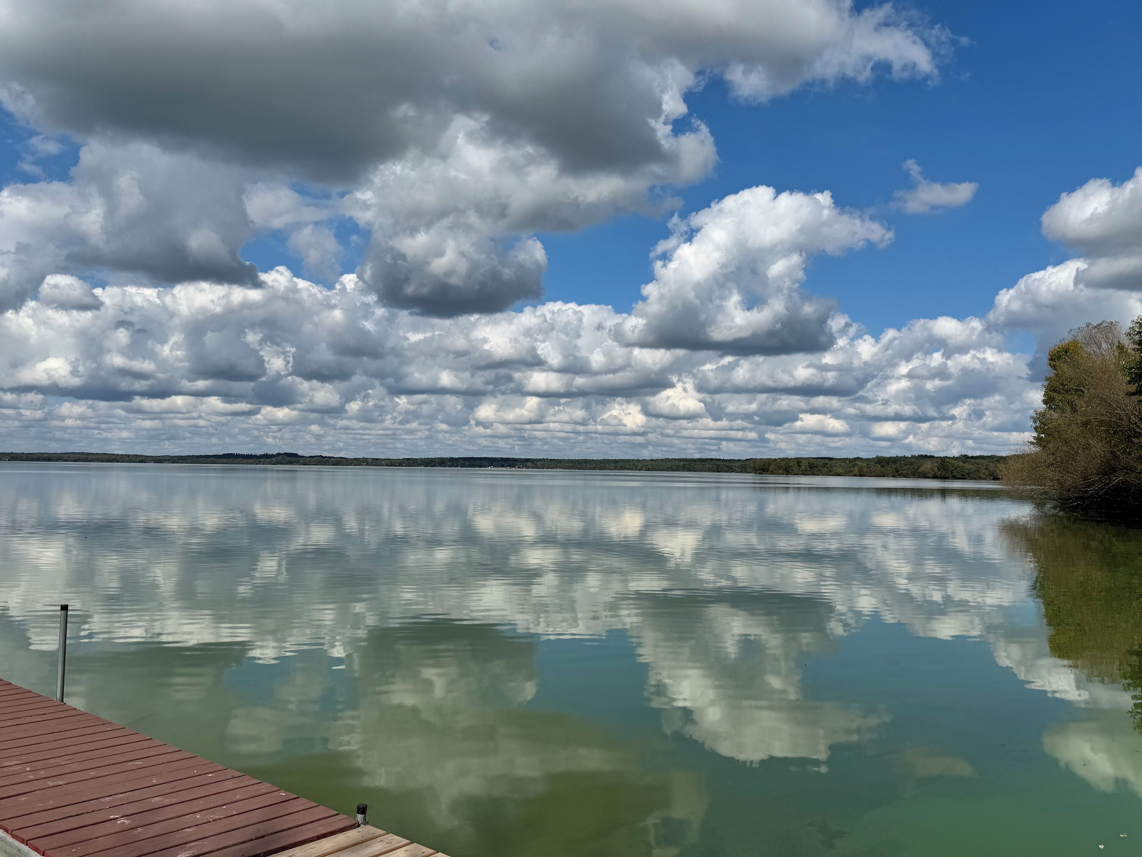 A calm lake with a wooden dock extending into the water, surrounded by trees and a cloudy sky.