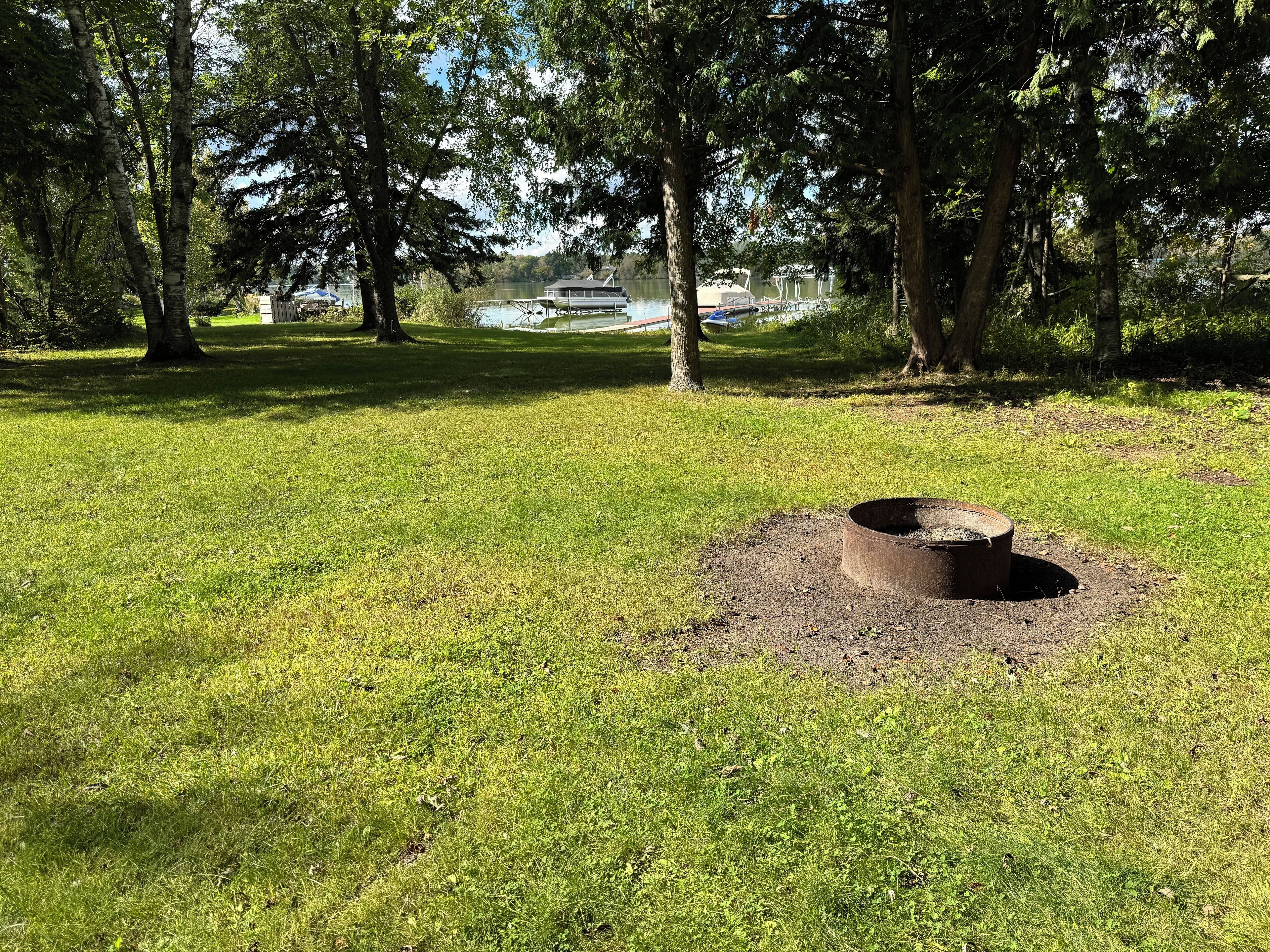 A grassy park area with a circular fire pit in the foreground and trees in the background.