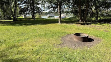 A grassy park area with a circular fire pit in the foreground and trees in the background.