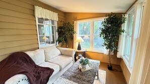 Ein gemütliches Zimmer mit einem beigen Sofa, einem kleinen Tisch mit einer Lampe und großen Fenstern mit Vorhängen.