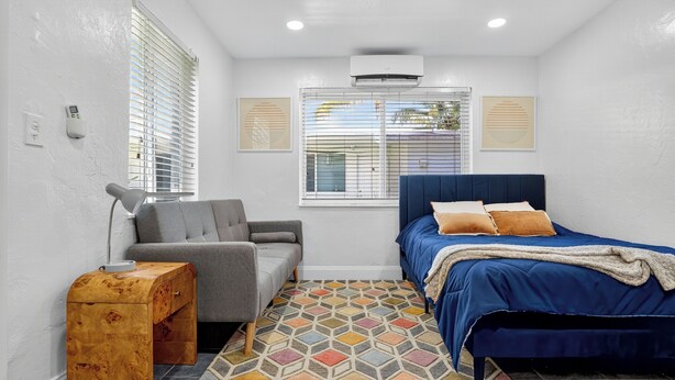 Une chambre à coucher comprenant un lit, une chaise, une petite table d’appoint en bois, un tapis à motifs et une fenêtre avec des stores.