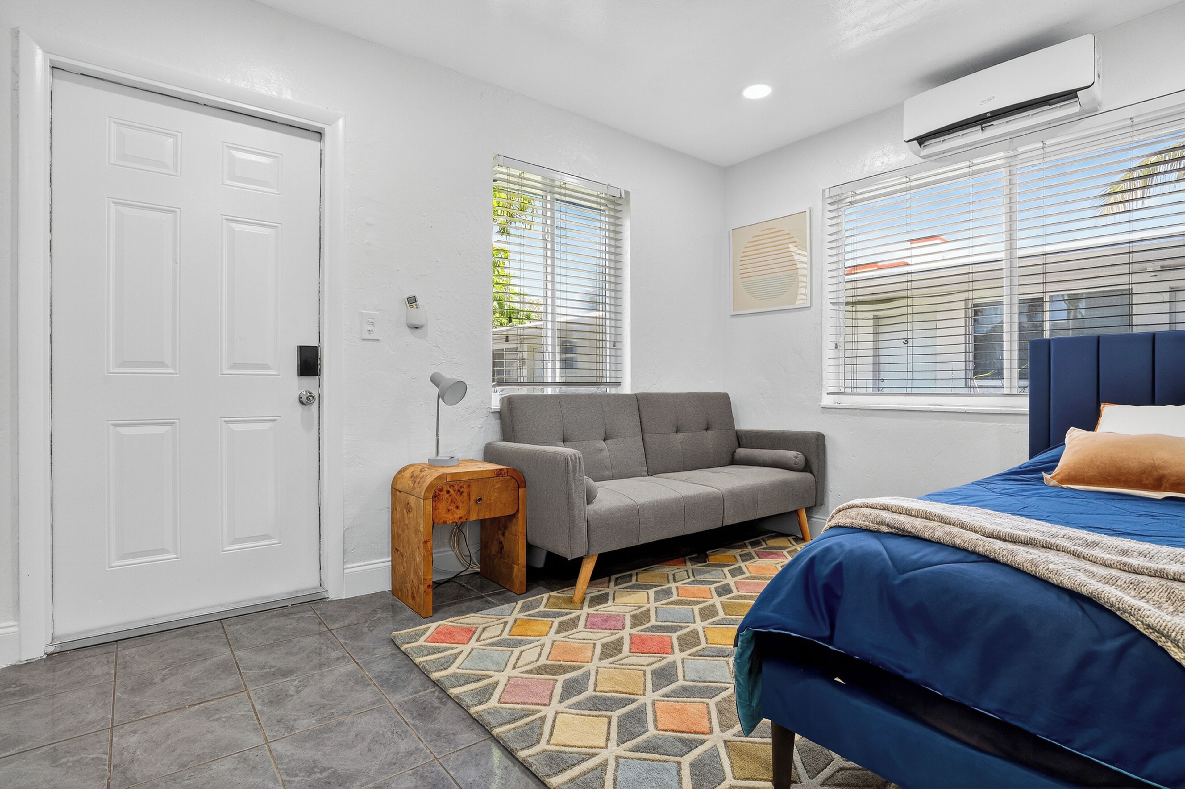 A bedroom with a bed, a grey sofa, a small wooden table, and a patterned rug.