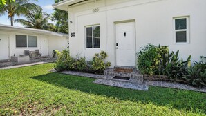 Une maison blanche à deux étages, avec le numéro « 60 » sur la porte d’entrée, un petit porche avec une table et des chaises, et une pelouse bien entretenue agrémentée de diverses plantes.