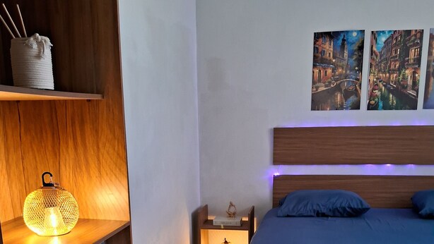 Een slaapkamer met een houten hoofdbord, een bed met blauw beddengoed, een nachtkastje met een lamp, een plank met handdoeken en een wanddecoratie.