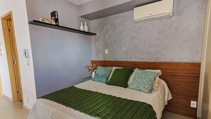 A bedroom with a bed, a wooden headboard, a grey wall, a shelf with decorative items, and an air conditioning unit. - Studio Pet Friendly c Garagem e Varanda no Centro (Juiz de Fora)