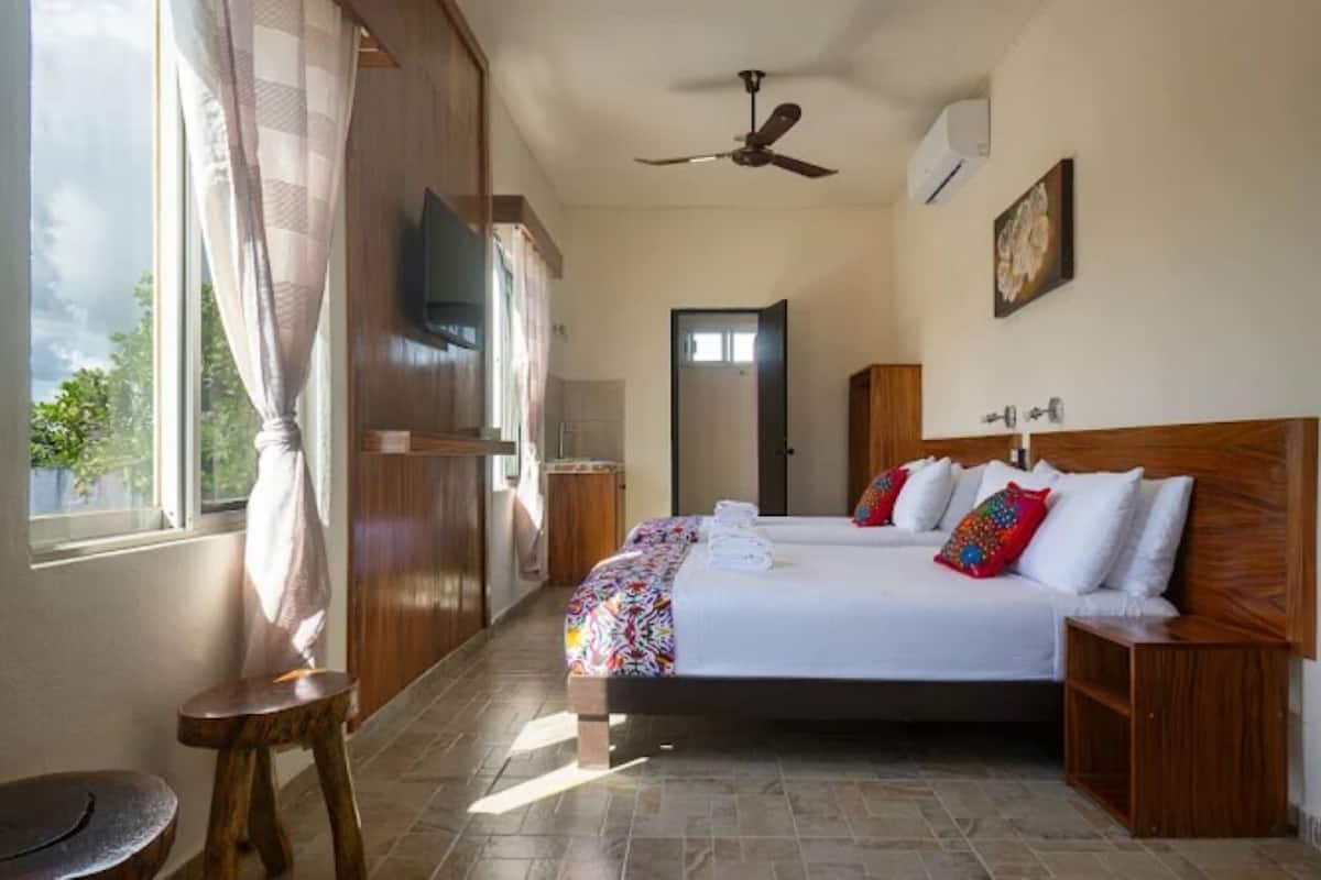 A hotel room with a bed, wooden headboard, a television, a ceiling fan, and a window with curtains.