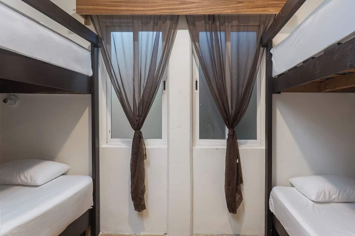 A bunk bed room with two beds, a wooden ceiling, and a window with sheer curtains.
