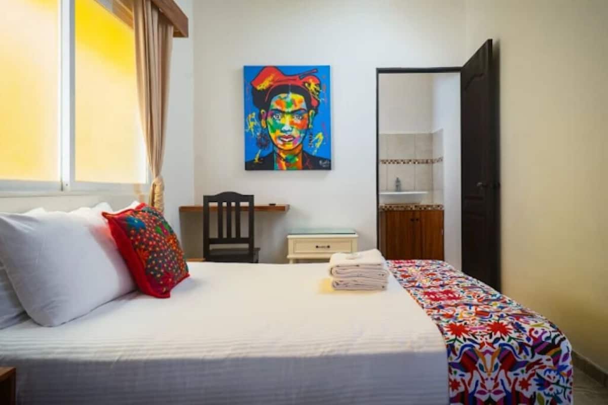 A hotel room with a bed, a colorful painting on the wall, a chair, a nightstand, and a door leading to another room.