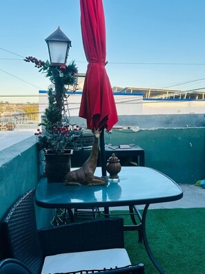 Uma área de pátio com mesa, cadeiras, um guarda-sol vermelho, uma girafa decorativa e um candeeiro.