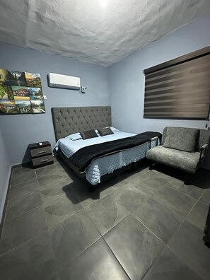 Quarto com cama, mesa de cabeceira, cadeira, ar condicionado fixado na parede e janela com persianas.