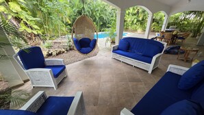 A covered outdoor seating area with wicker furniture and blue cushions, surrounded by lush greenery and a pool. - 3-bedroom villa in beautiful Sosúa with cool AC breeze (Sosúa)