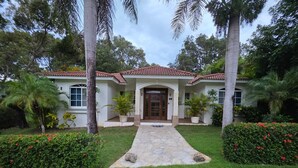 A two-story house with a red-tiled roof, a front entrance with a wooden door, and a stone pathway leading to the door. - 3-bedroom villa in beautiful Sosúa with cool AC breeze (Sosúa)
