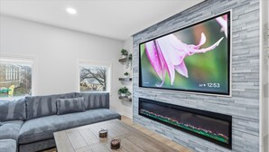 A modern living room with a grey sofa, a wooden coffee table, a flat-screen TV mounted on a grey brick wall, and a fireplace below the TV. - Amazing remodeled property in the Heart of Indy (Indianapolis)