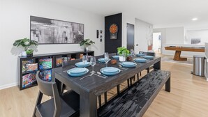 A modern dining area with a long table set for six, surrounded by black chairs. There are two potted plants on the table, and a dartboard is mounted on the wall. - Amazing remodeled property in the Heart of Indy (Indianapolis)