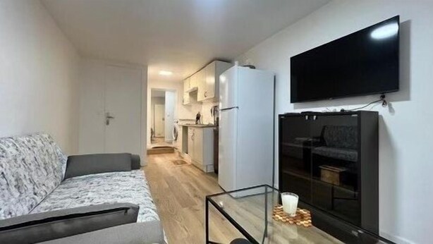 Un espace de vie compact comprenant un canapé, une table basse avec un plateau en verre, un téléviseur à écran plat fixé au mur et, au fond, une petite cuisine.