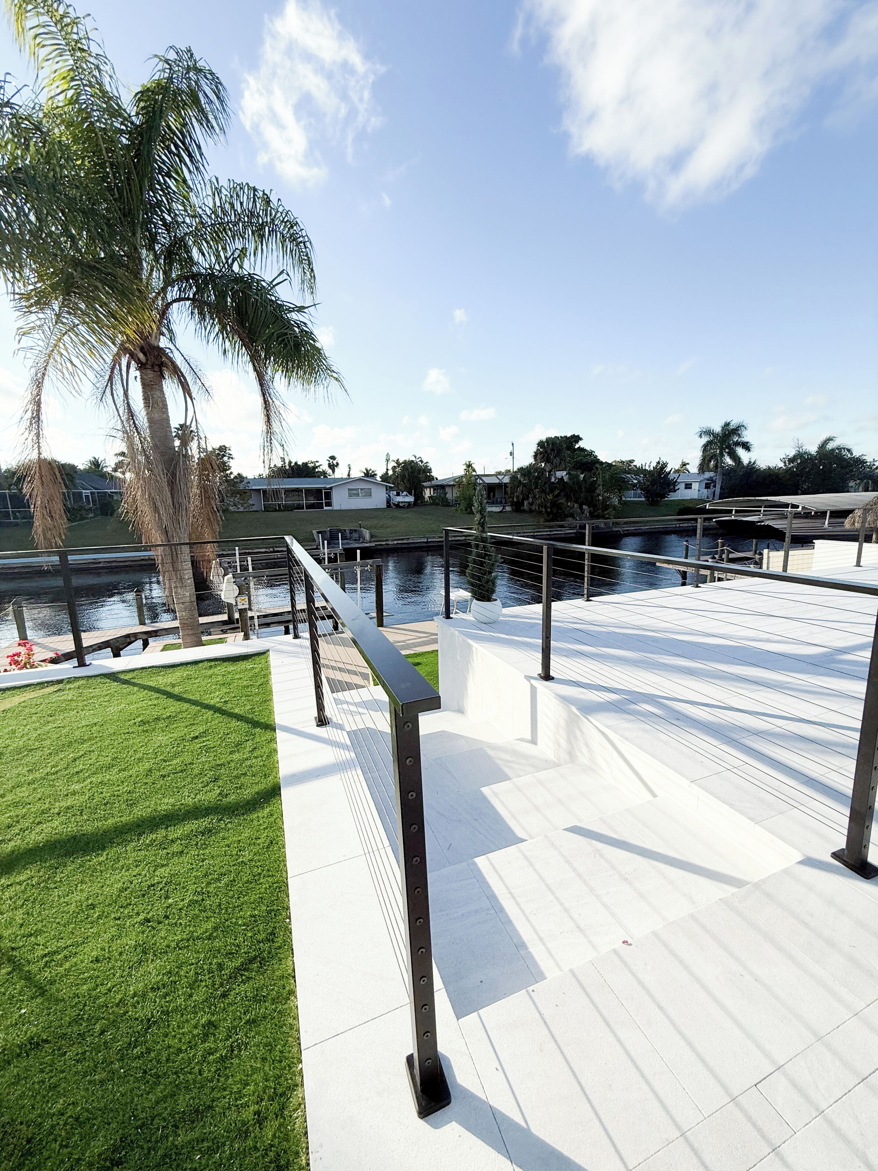 A waterfront promenade with a metal railing, a palm tree, and a view of the water and buildings in the background.