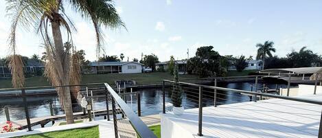 A waterfront promenade with a metal railing, a palm tree, and a view of the water and buildings in the background.