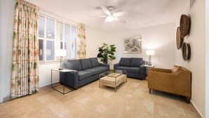Ein Wohnzimmer mit zwei grauen Sofas, einem Holztisch, einem braunen Sofa, einem Deckenventilator und einem großen Fenster mit Vorhängen.