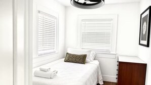 A bedroom with a bed, two windows with blinds, a bedside table, and a wooden cabinet. - Relax in this cozy home near downtown & stadiums with everything you need. (Houston)