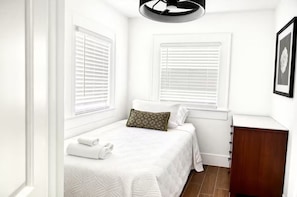 A bedroom with a bed, two windows with blinds, a bedside table, and a wooden cabinet. - Relax in this cozy home near downtown & stadiums with everything you need. (Houston)