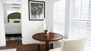 A hotel room with a bed, a round wooden table with a chair, a framed picture on the wall, and a window with blinds. - Relax in this cozy home near downtown & stadiums with everything you need. (Houston)