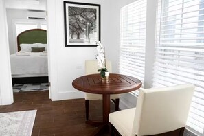 A hotel room with a bed, a round wooden table with a chair, a framed picture on the wall, and a window with blinds. - Relax in this cozy home near downtown & stadiums with everything you need. (Houston)